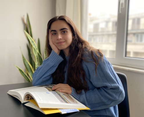 A portrait of Katerina sitting at a desk with an open book, resting her head on her hand in a brightly lit room.