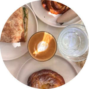 A top-down view of a round table featuring a latte with latte art, a glass of water, a pastry, and a sandwich cut in half.