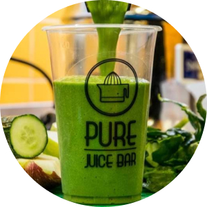 A clear plastic cup with the "Pure Juice Bar" logo being filled with a bright green juice, surrounded by fresh ingredients like cucumber slices and leafy greens.