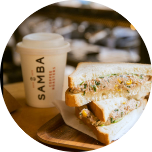 A stacked sandwich cut in half filled with a savory filling, sitting next to a white paper coffee cup with the "Samba Coffee Roasters" logo on a wooden surface.
