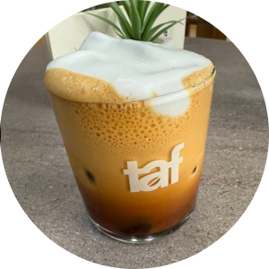 A tall glass filled with a layered iced coffee drink topped with a thick, frothy milk foam, featuring the "taf" logo on the glass